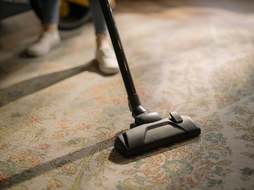 A yellow vacuum cleaner with a flexible hose is positioned on a patterned area rug in a living room, adjacent to a wooden sideboard. The sideboard holds a chessboard, a white sculptural bust, a remote