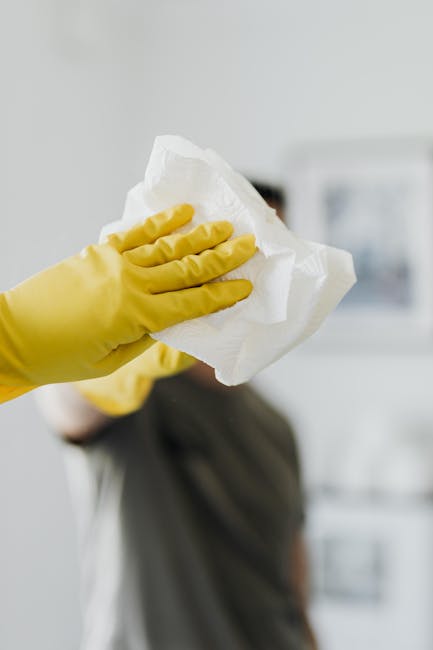 A person wearing yellow rubber gloves is holding a crumpled white paper towel, preparing for surface cleaning. In the background, a blurred kitchen setting features a stainless steel appliance and lig