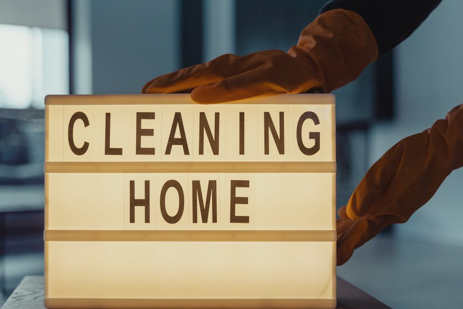 A person wearing yellow rubber gloves is holding a crumpled white paper towel, preparing for surface cleaning. In the background, a blurred kitchen setting features a stainless steel appliance and lig